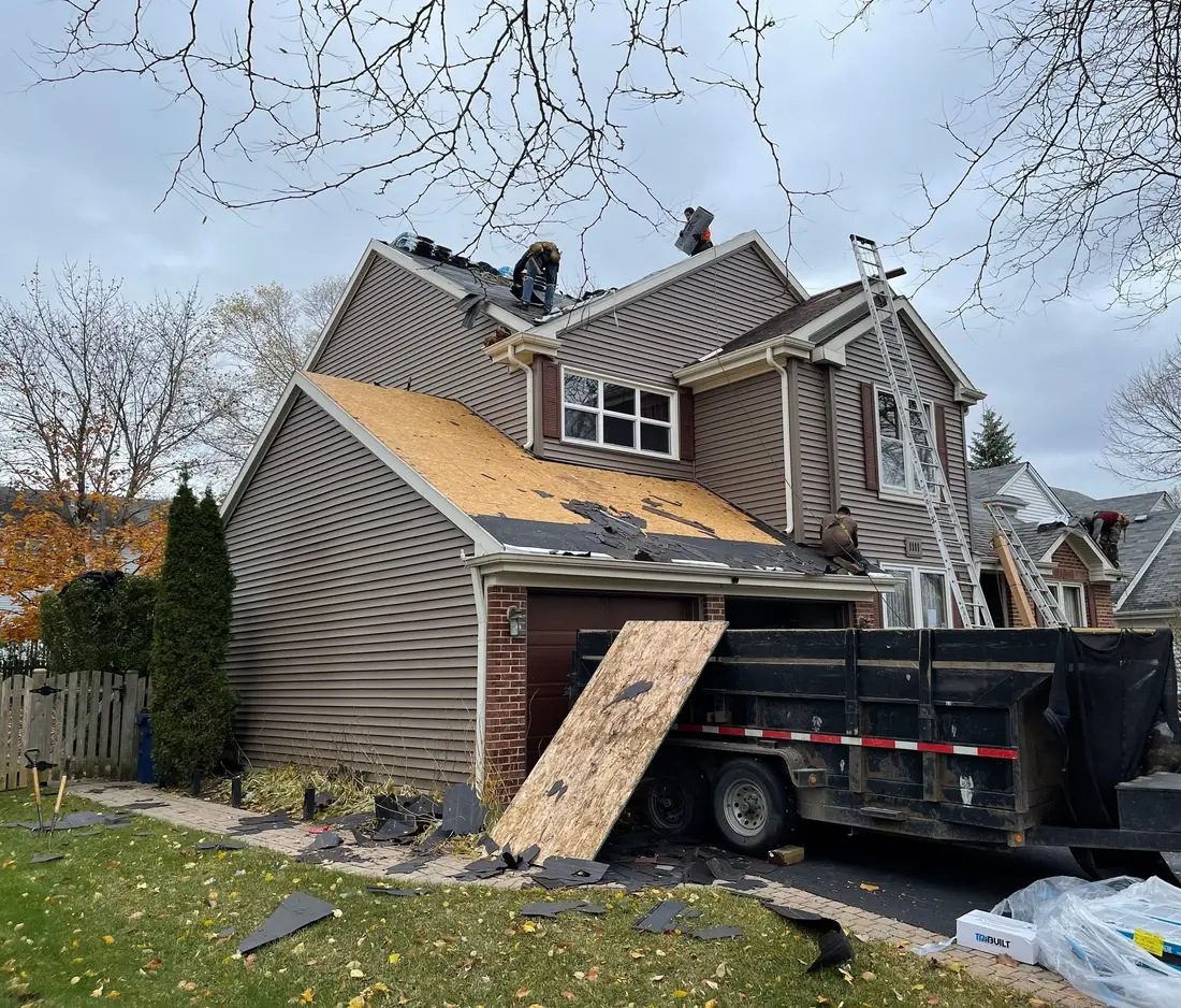 Side of a two-story house with half of the old roof stripped, a sheet of plywood leaning on a dump trailer in the driveway
