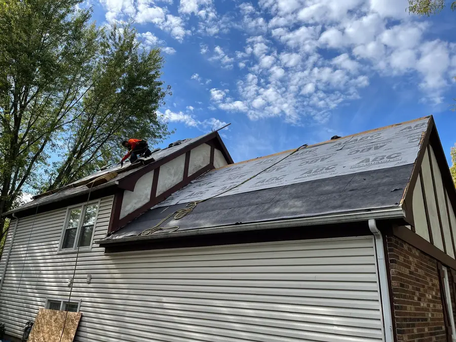 Roofer working on the slope beside a stucco dormer, with old shingles partly stripped and white vinyl siding below