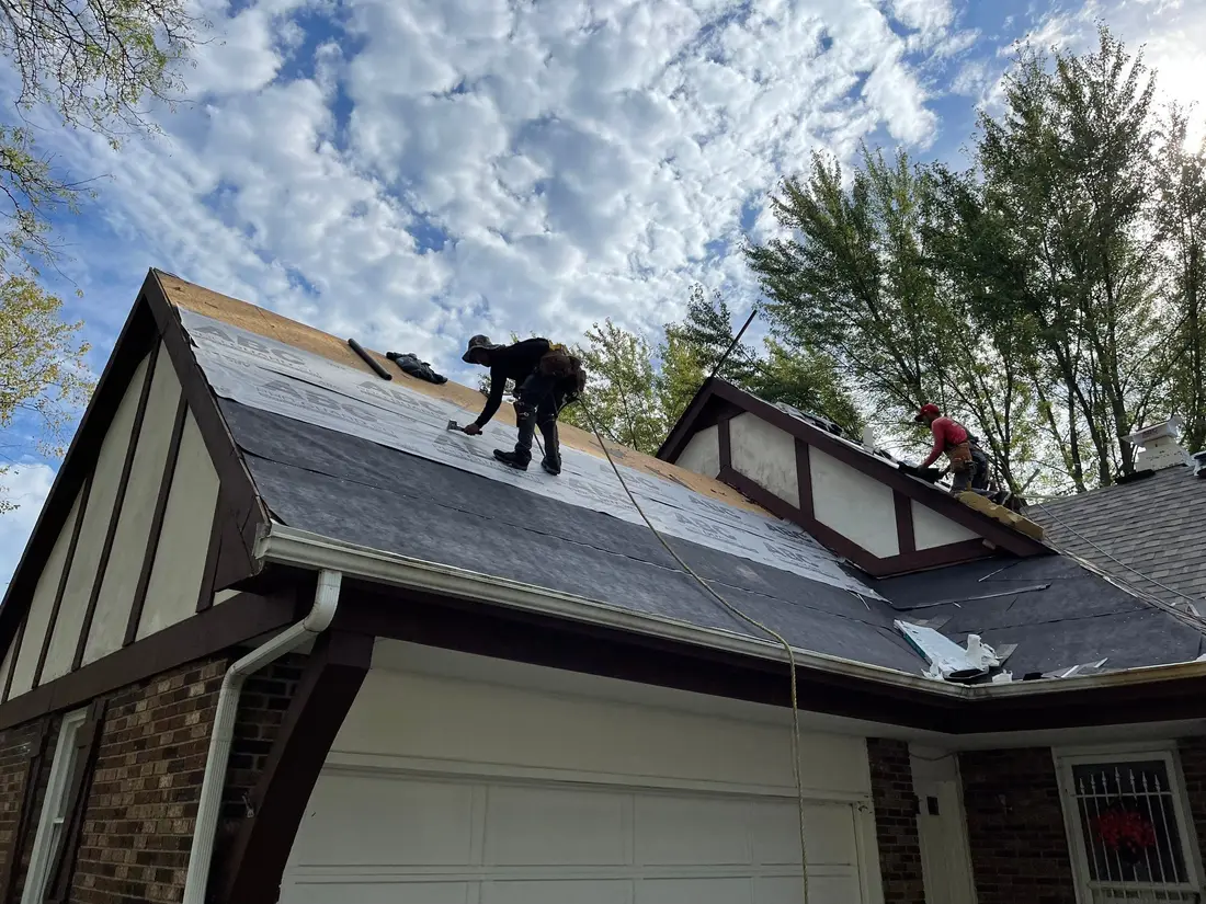 Two roofers installing underlayment on a steep gable of a brick and stucco house under a blue sky