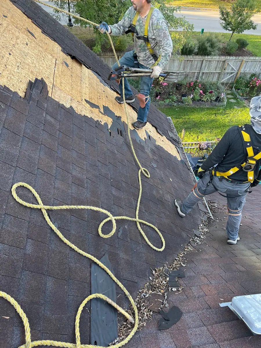 Roofer in a yellow safety harness working on a slope during a tear-off, with a rope coiled on the old shingles