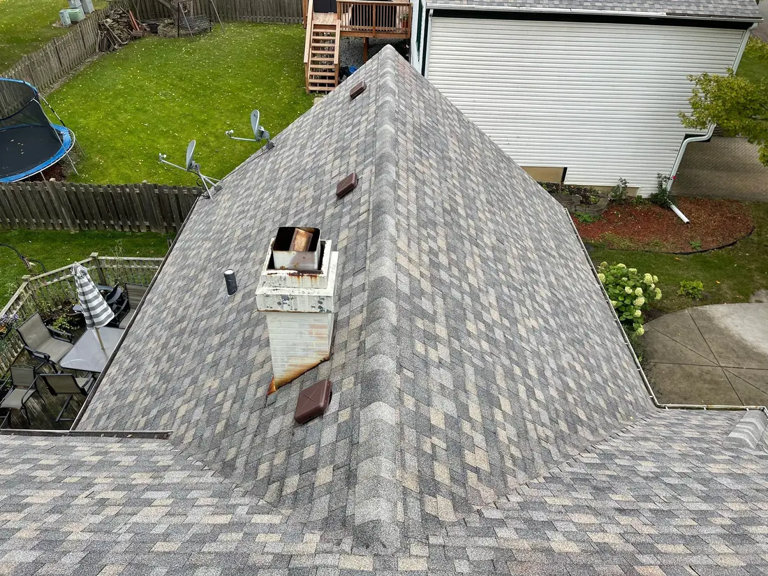 New gray architectural shingles meeting at a ridge beside a white chimney chase, with a backyard below