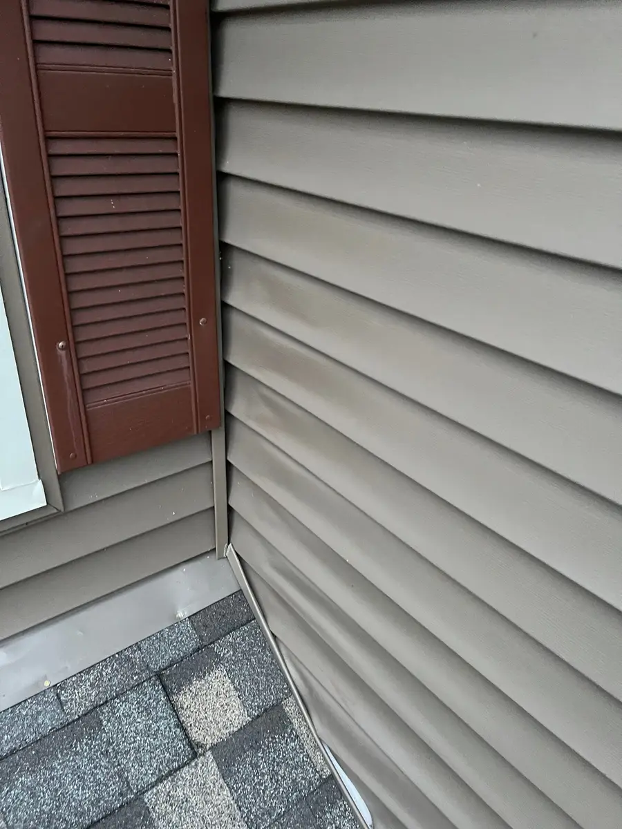 Tan vinyl siding with a brown louvered shutter at an inside corner above a roof line