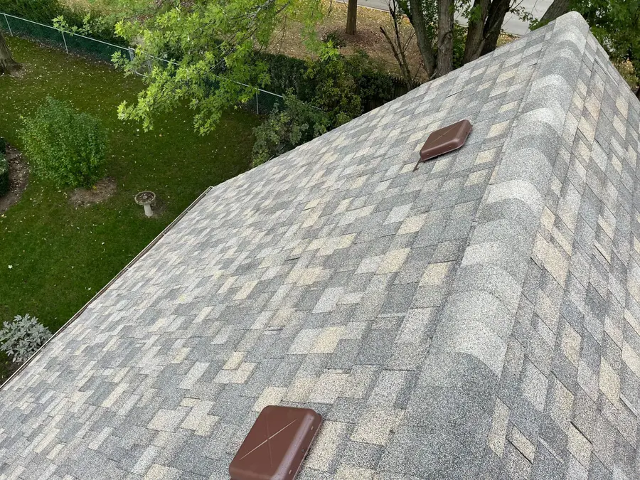 Freshly shingled slope with a brown box vent near the ridge above a garden
