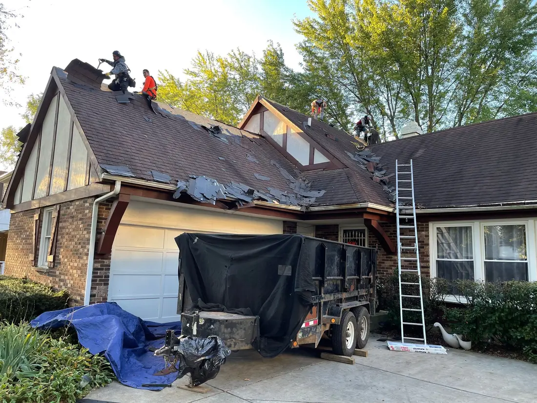 Dump trailer covered with a black tarp in a driveway while roofers strip the shingles from a brick and stucco house