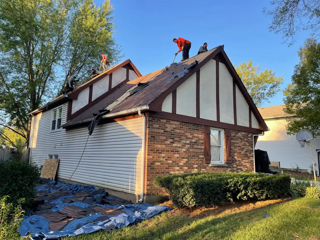 Roofers stripping old shingles from a steep gable, with tarps spread on the lawn below